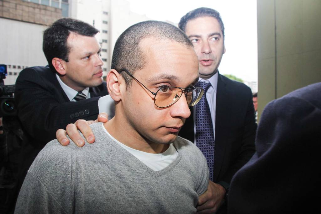 Imagem mostra Gil Rugai cercado por advogado e jornalista durante depoimento. Ele veste camisa branca, blusa cinza e utiliza óculos de grau na cor laranja