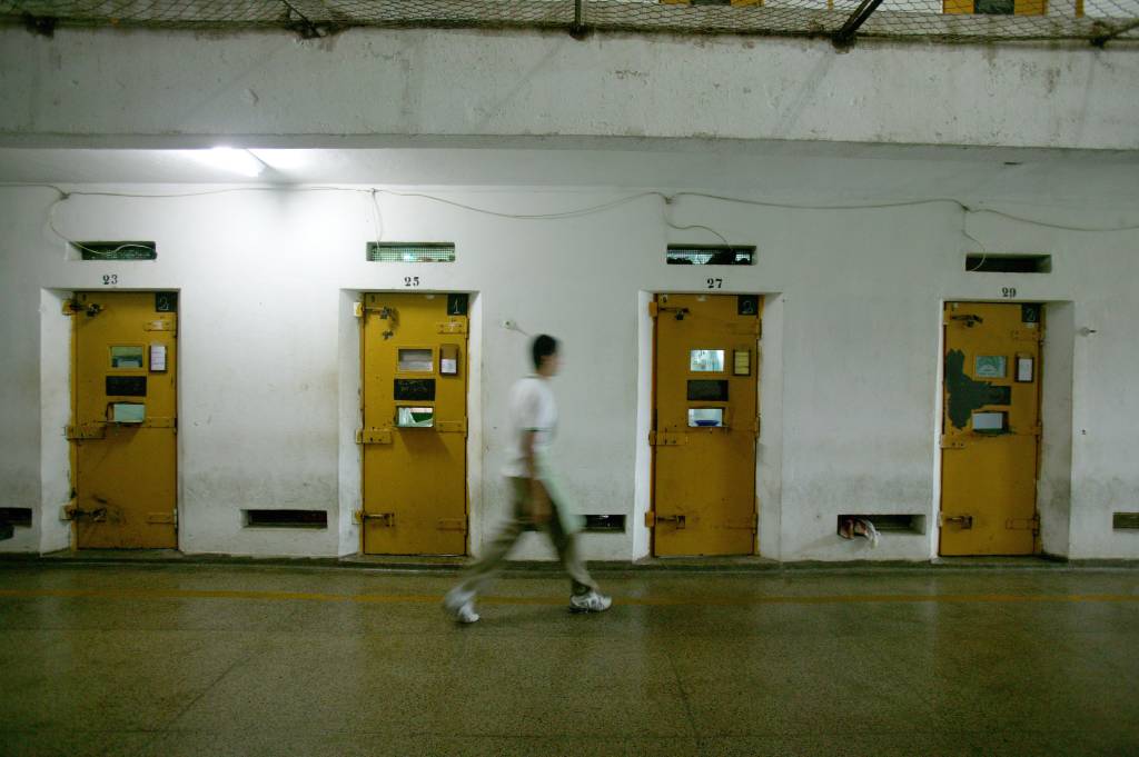 A imagem mostra um preso andando por corredores de uma penitenciária