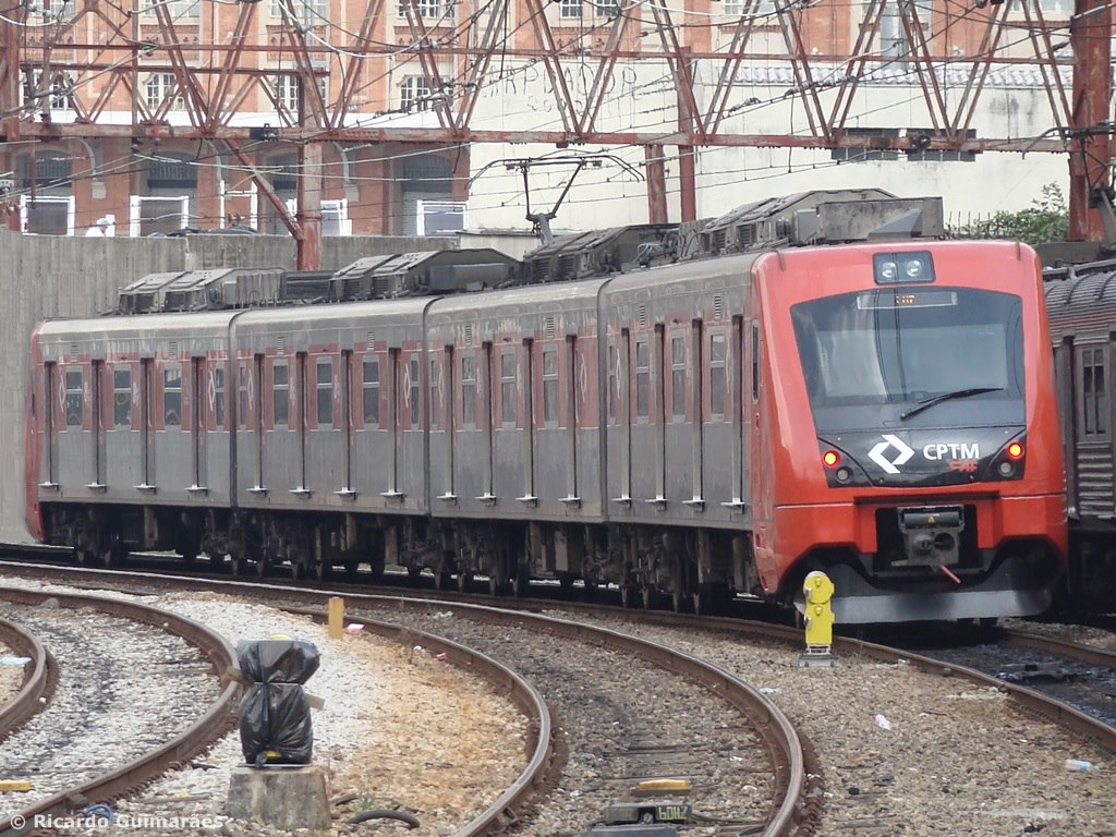 Imagem mostra trem se movendo em trilhos. Ao fundo, uma passarela passa por cima do trem