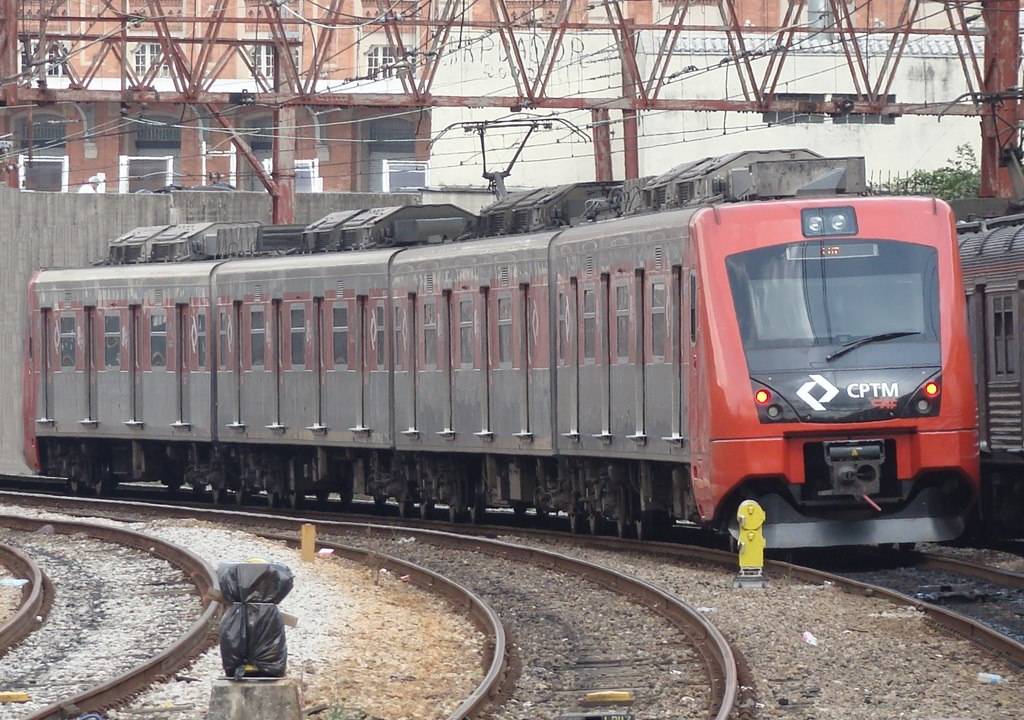 Imagem mostra trem se movendo em trilhos. Ao fundo, uma passarela passa por cima do trem