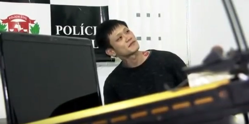 Imagem mostra Denis sentado em delegacia de polícia, usando camiseta, olhando para a direita
