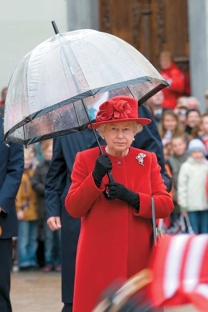 A imagem mostra a rainha Elizabeth de vestido na rua segurando um guarda-chuva