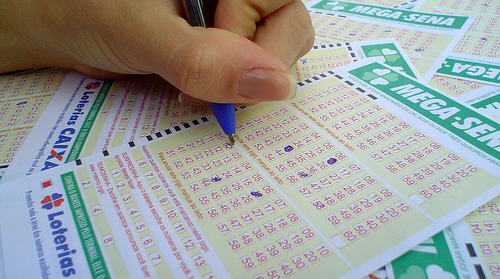 A imagem mostra uma mão com uma caneta preenchendo quadradinhos de uma aposta em loteria.