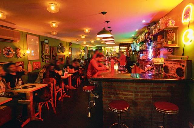 Balcão de bar com mesas à esquerda. Luzes coloridas