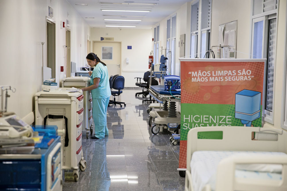imagem mostra corredor de hospital, com camas e diversos itens hospitalares no meio do caminho e uma mulher em pé, com roupas hospitalares