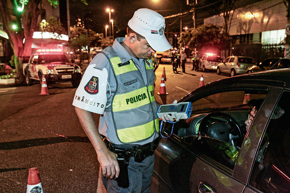 A imagem mostra um policial militar, ao lado da porta do motorista de um carro, com um bafômetro na mão.