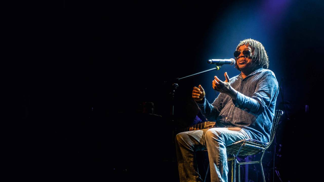 Imagem mostra homem sentado com os braços levantados em palco, cantando em microfone