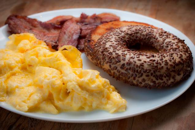 O café da manhã é composto de bagel na chapa, ovos mexidos e bacon, e vem com um potinho de cream cheese