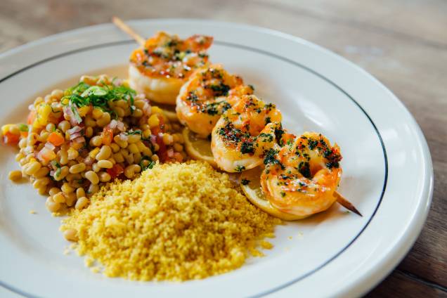 Espetinho de camarão: feito na brasa, acompanha salada de feijão-manteiguinha e um mix de farinhas de milho, pão e biscoito de polvilho Espetinho de camarão: feito na brasa, acompanha salada de feijão-manteiguinha e um mix de farinhas de milho, pão e biscoito de polvilho