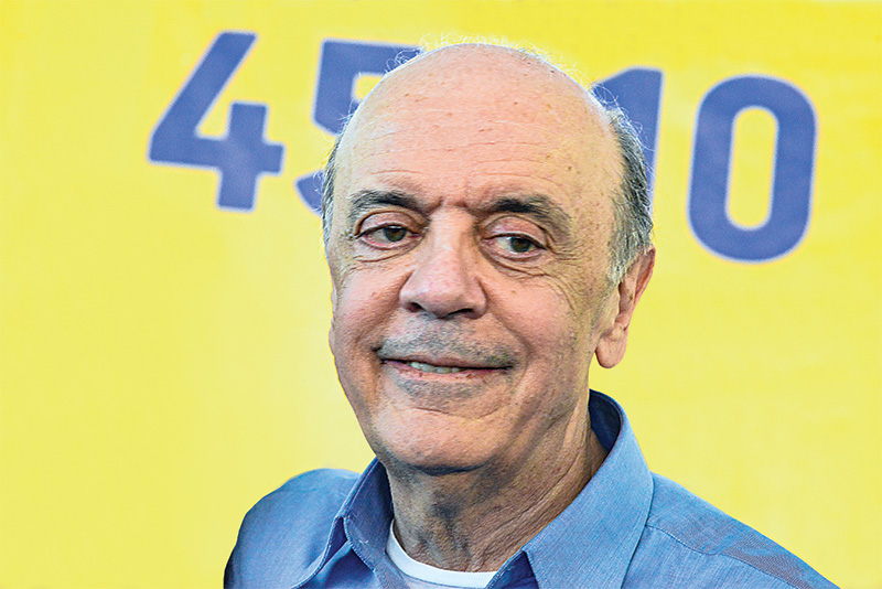 Imagem mostra José Serra sorrindo, sob fundo amarelo, com números em azul
