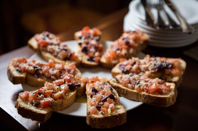 Montadas sobre um pão macio, a porção de enormes bruschettas traz tomate picado, azeitona preta e parmesão