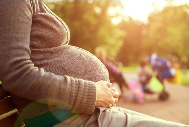 A imagem mostra a barriga de uma mulher grávida; ela está sentada em um banco de praça