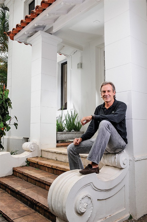 Homem branco grisalho e cacreca de roupas pretas posa em frente a mesa com livro grande. Atrás, prateleiras exibem mais livros