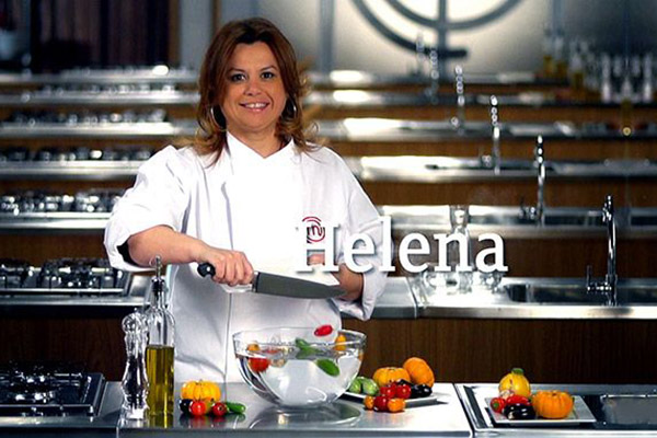 Helena Manosso sorrindo atrás de bancada do MasterChef enquanto despeja legumes cortados de tábua para tigela de vidro.