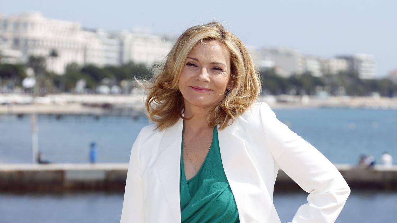 Kim Cattrall posa em frente ao mar com camiseta verde e blazer branco.