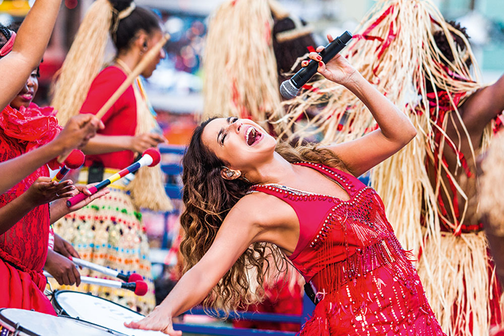 Foto de Daniela Mercury em show, de vestido vermelho, com microfone na mão