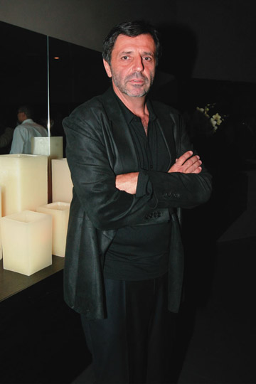 Homem branco de cabelos castanhos curtos dá risada olhando à direita em frente a parede com mapa múndi preto. Veste camiseta preta.