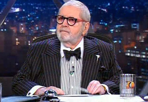 Foto de Jô Soares usando terno, gravata e óculos, sentado em sua bancada no Programa do Jô