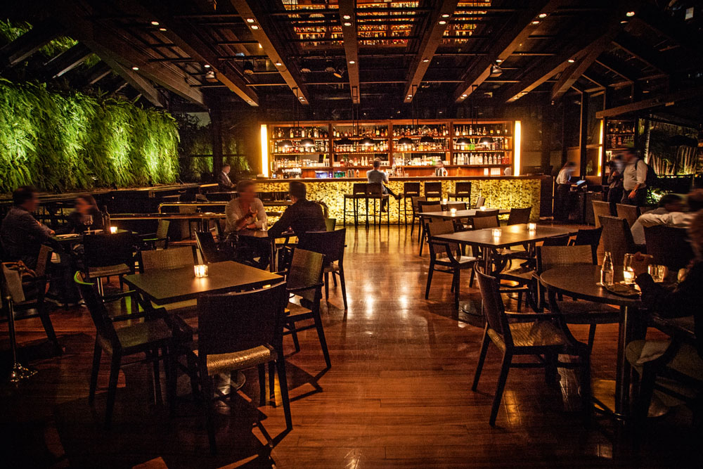 Foto exibe mesas, chão de madeira escura e balcão de bar ao fundo