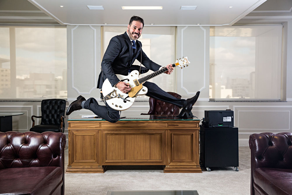 Imagem mostra Fernando Parrillo, de terno e gravata, pulando com guitarra os braços em escritório empresarial
