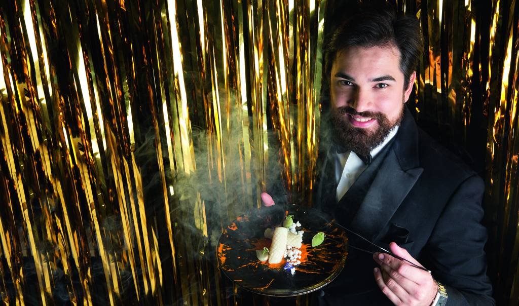 Chef Rodrigo Aguiar posa em frente a fundo dourado enquanto segura um prato