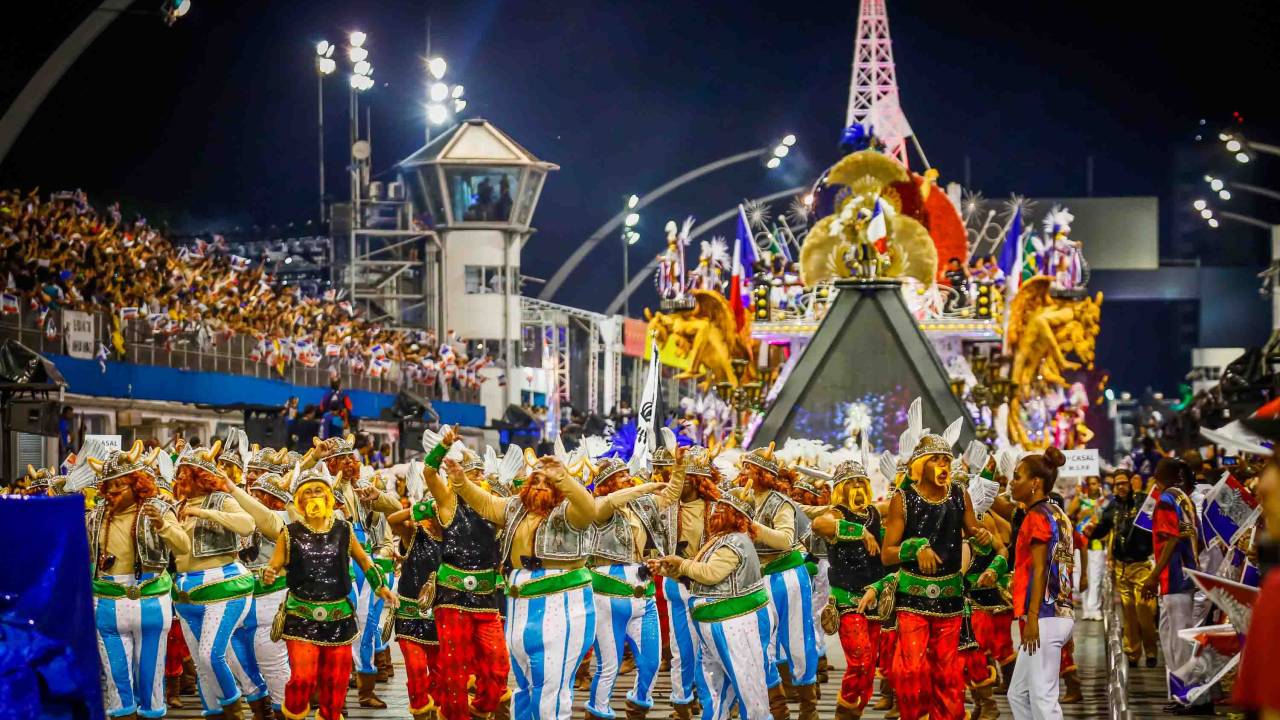 Imagem mostra desfile de escola de samba no Anhembi, com pista e arquibancada lotadas e carro alegórico ao fundo