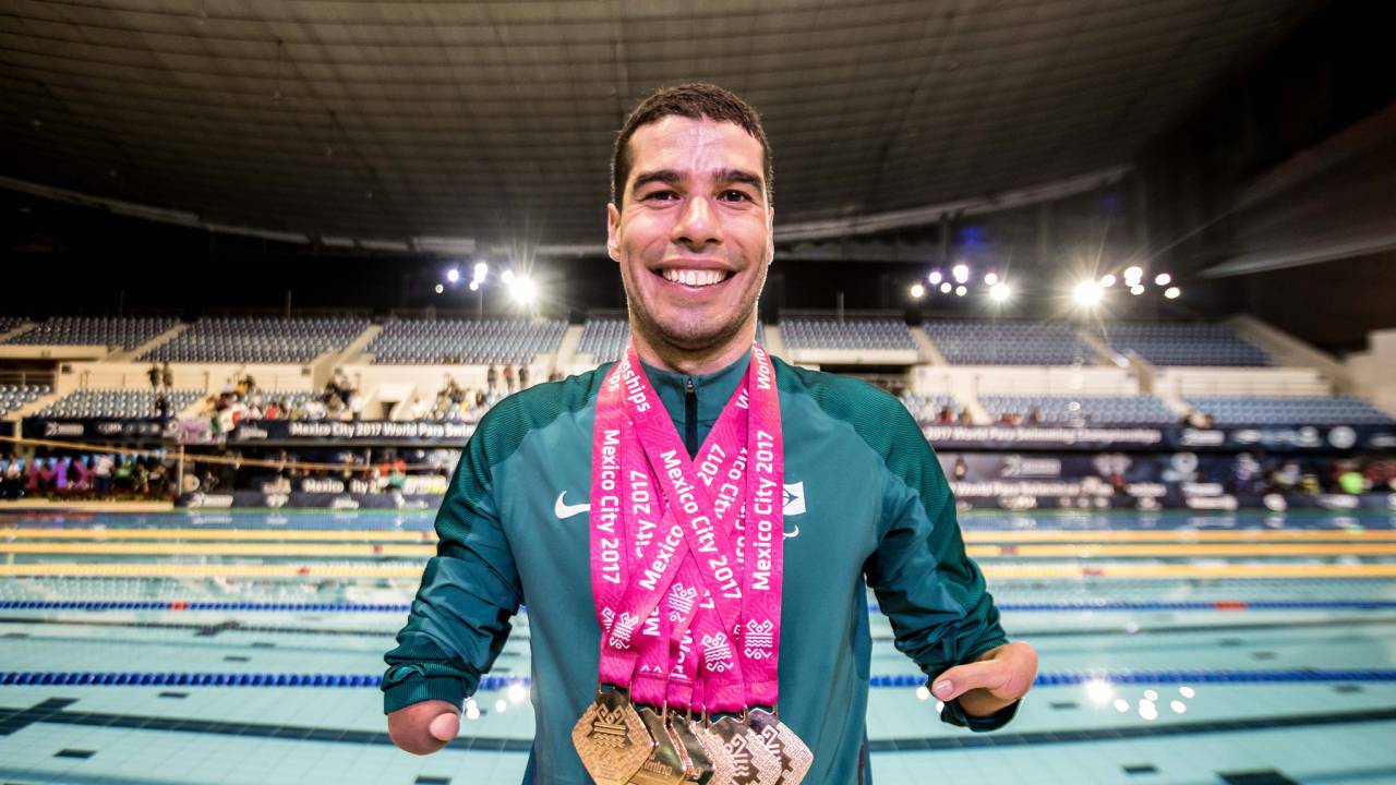 Imagem mostra Daniel Dias com diversas medalhas no pescoço. Ele está na frente de piscina olímpica