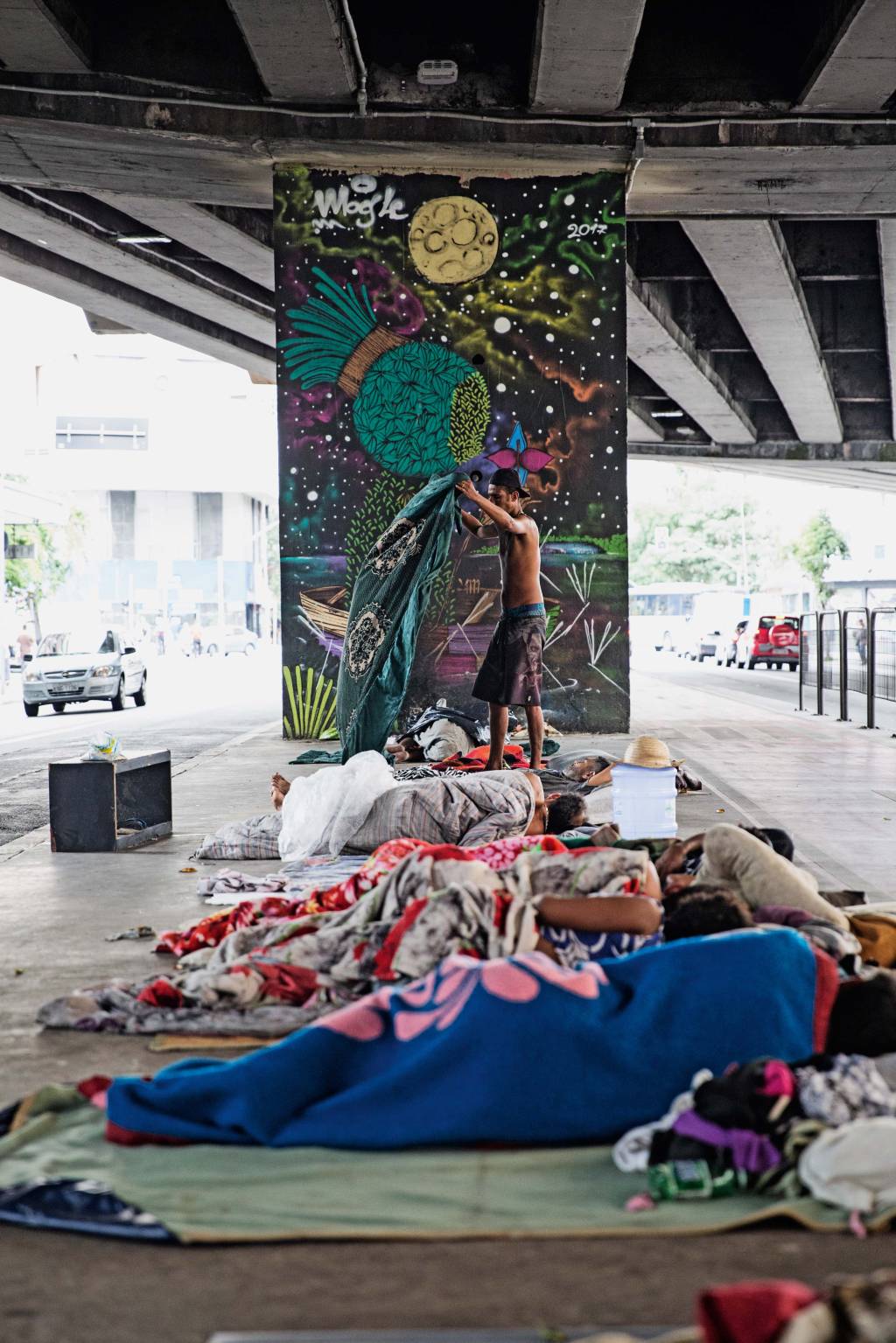 Moradores de rua deitados debaixo do Minhocão. No centro da foto, homem estica cobertor.