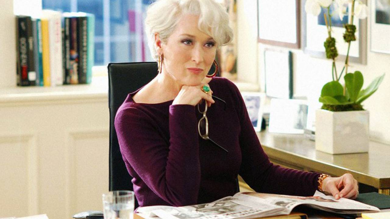 Meryl Streep em O Diabo Veste Prada aparece com uma mão no queixo analisando alguém. Segura os óculos de grau, tem cabelos brancos e veste suéter roxo. Está sentada na mesa com revista aberta.