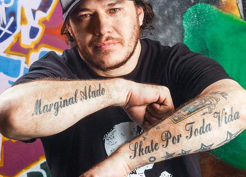 Chorão mostra os braços tatuados. Usa boné e camiseta preta com caveira de estampa.