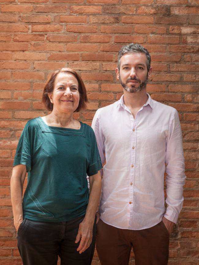 Isabel e Gonçalo posam de pé lado a lado. Ela veste camiseta verde e calça preta. Ele veste camisa rosada e calça escura. Ao fundo, tijolos aparecem na foto.