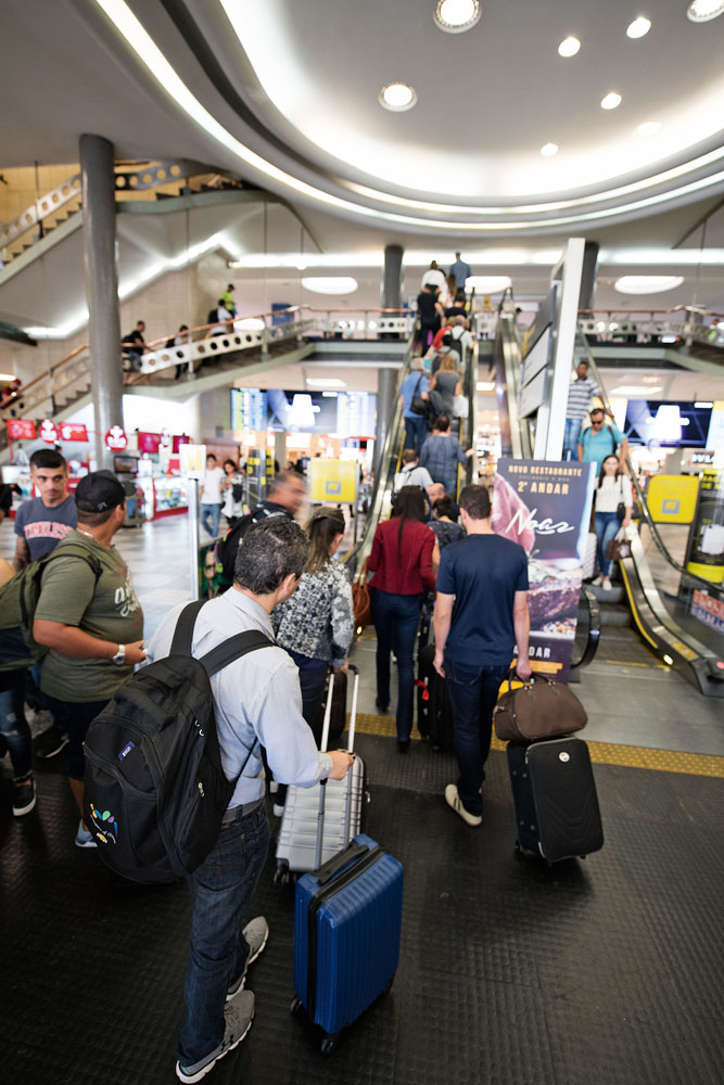 Foto mostra passageiros com malas andando em direção a escada rolante