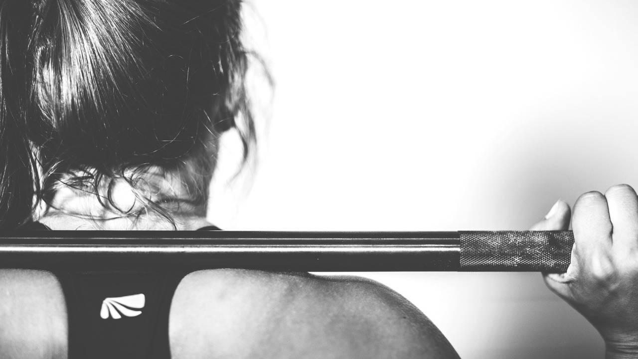 Imagem mostra mulher, de costas, com barra de ferro apoiada nas costas, fazendo exercício