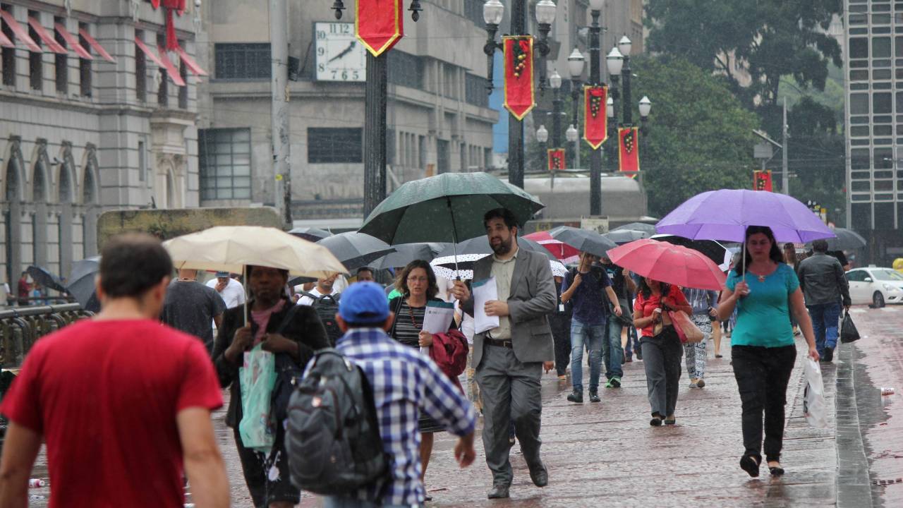 A imagem mostra pessoas caminhando em meio à chuva