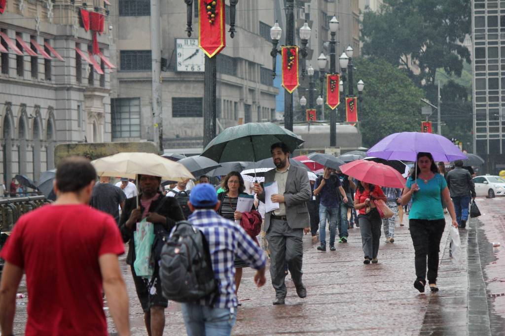 A imagem mostra pessoas caminhando em meio à chuva