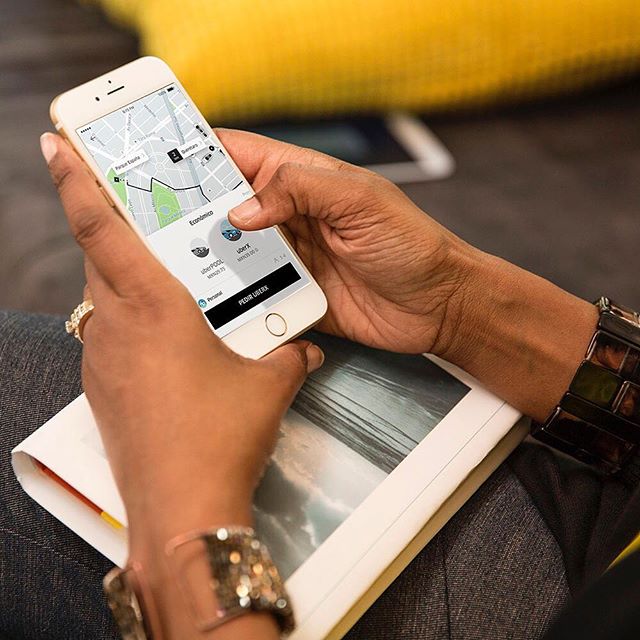 Imagem mostra mulher com celular na mão, acionando viagem pelo aplicativo da Uber. Ela segura também um livro e usa pulseiras no braço esquerdo, além de anéis nos dedos