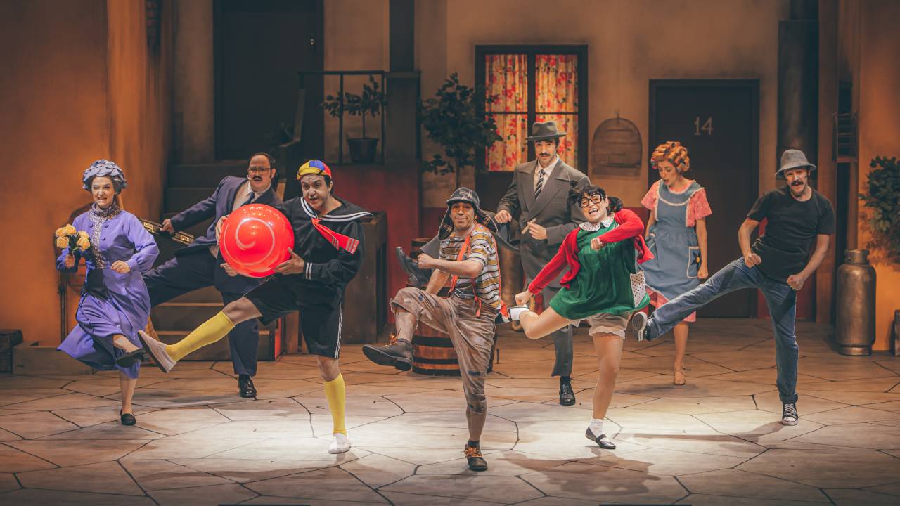 Foto exibe cena de espetáculo musical do Chaves com todo o elenco dançando em cenário inspirado na vila do seriado.