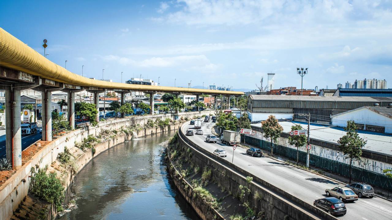 Imagem mostra Rio Tamanduateí ao centro da imagem. Ao lado direito, pista da Avenida do Estado, com poucos veículos em dia de sol