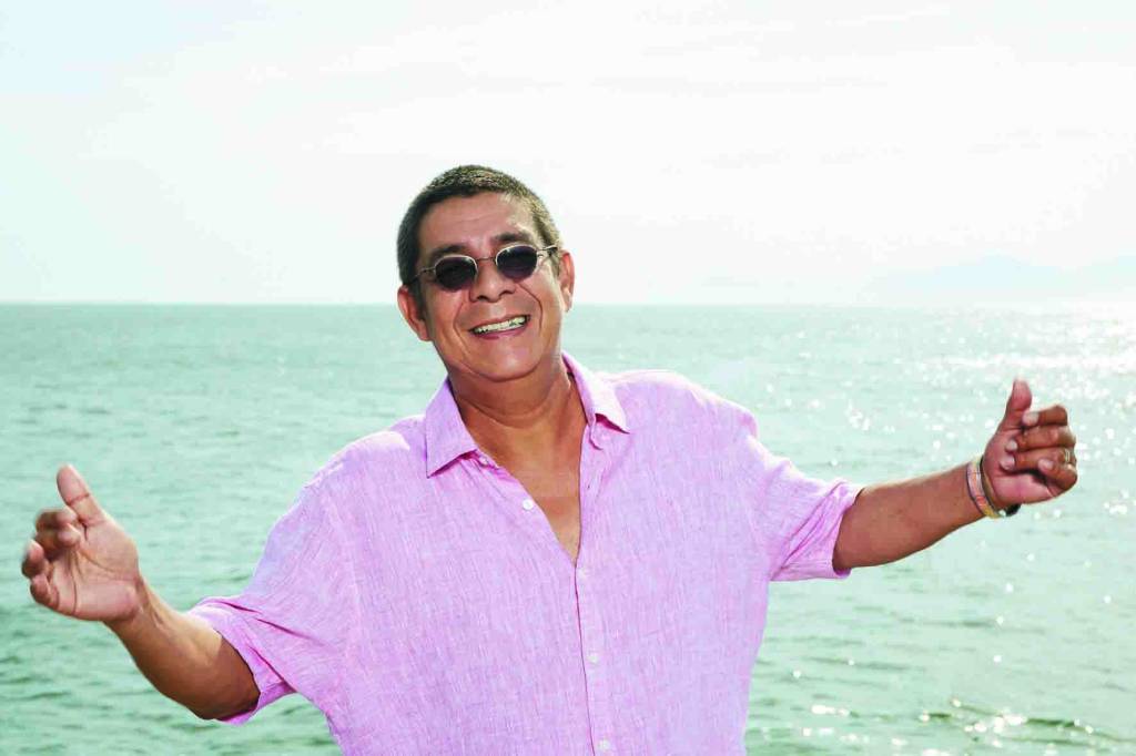 Imagem mostra Zeca Pagodinho sorrindo, usando óculos, na frente do mar