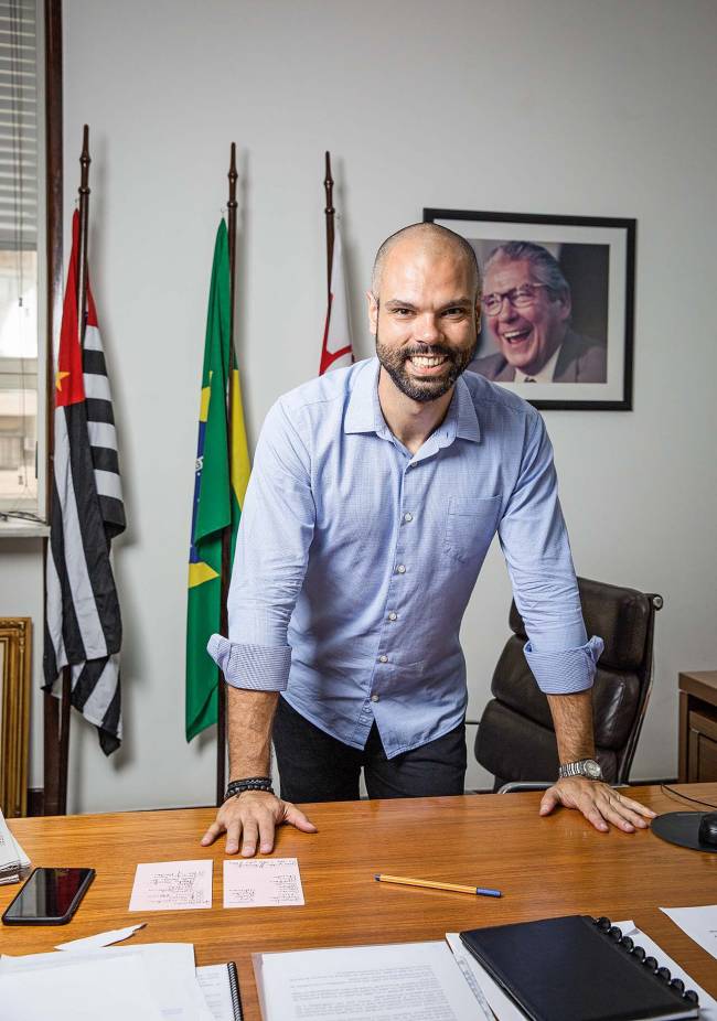 A imagem mostra Bruno Covas, em seu gabinete, de pé, com duas mãos sobre sua mesa e sorrindo para a câmera