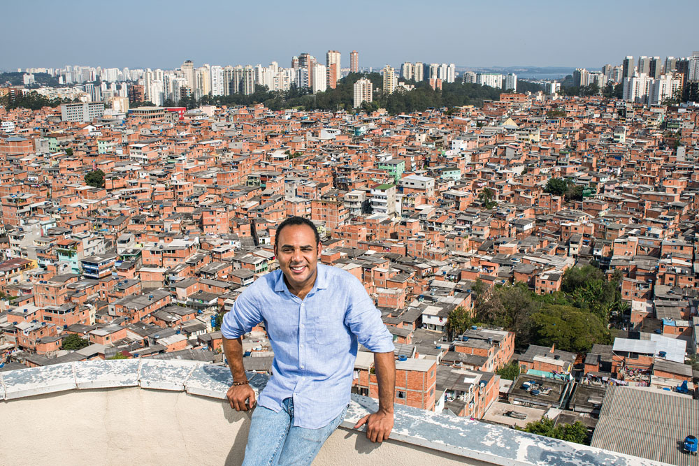 Gilson aparece sentado em muro de concreto com comunidade de Paraisópolis ao fundo. veste jeans clara e camisa azul claro.