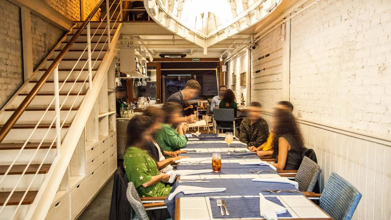 Parte térrea do salão do restaurante Lido Amici di Amici com pessoas sentadas em uma mesa coletiva