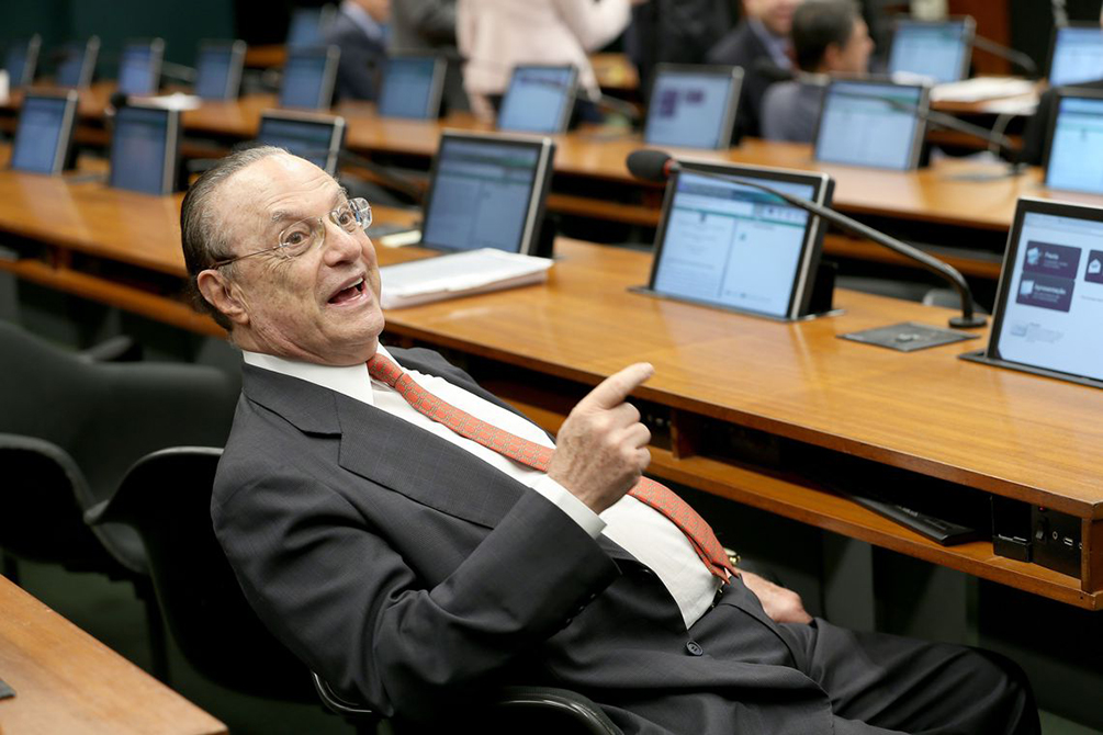 O ex-deputado federal Paulo Maluf aparece sentado em cadeira e em frente a balcão com telas de computador.
