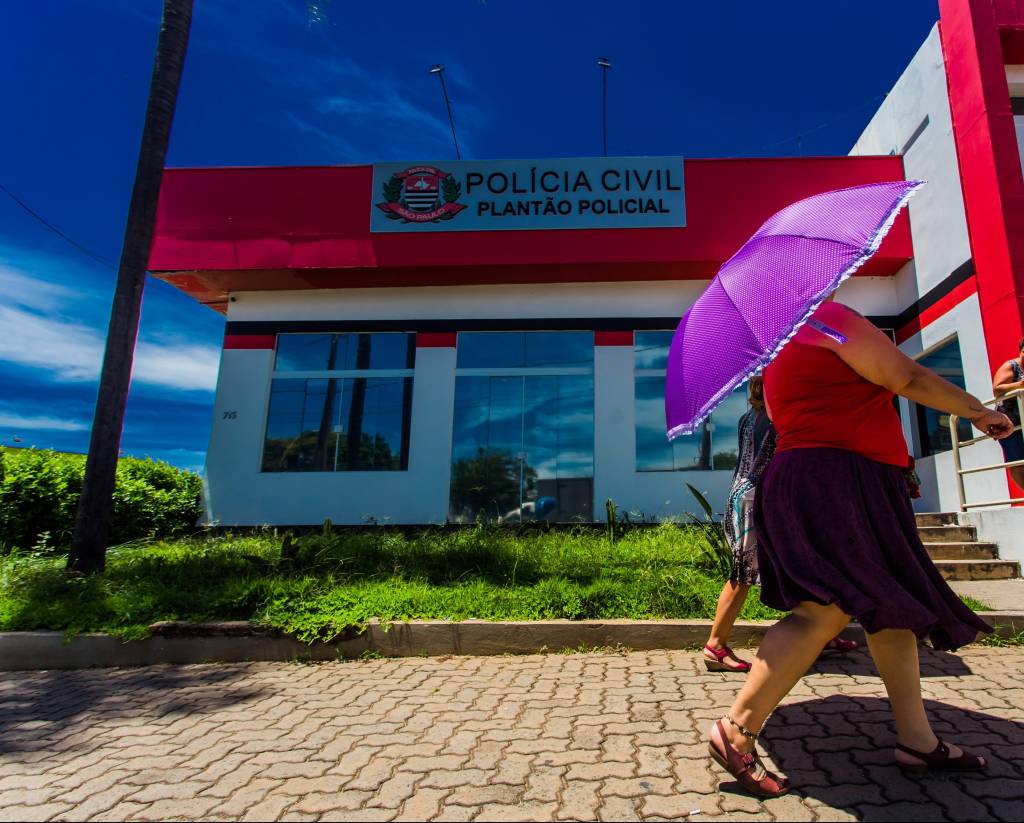 Fachada de unidade de Delegacia de Defesa da Mulher com pedestre segurando guarda-chuva rosa à direita.