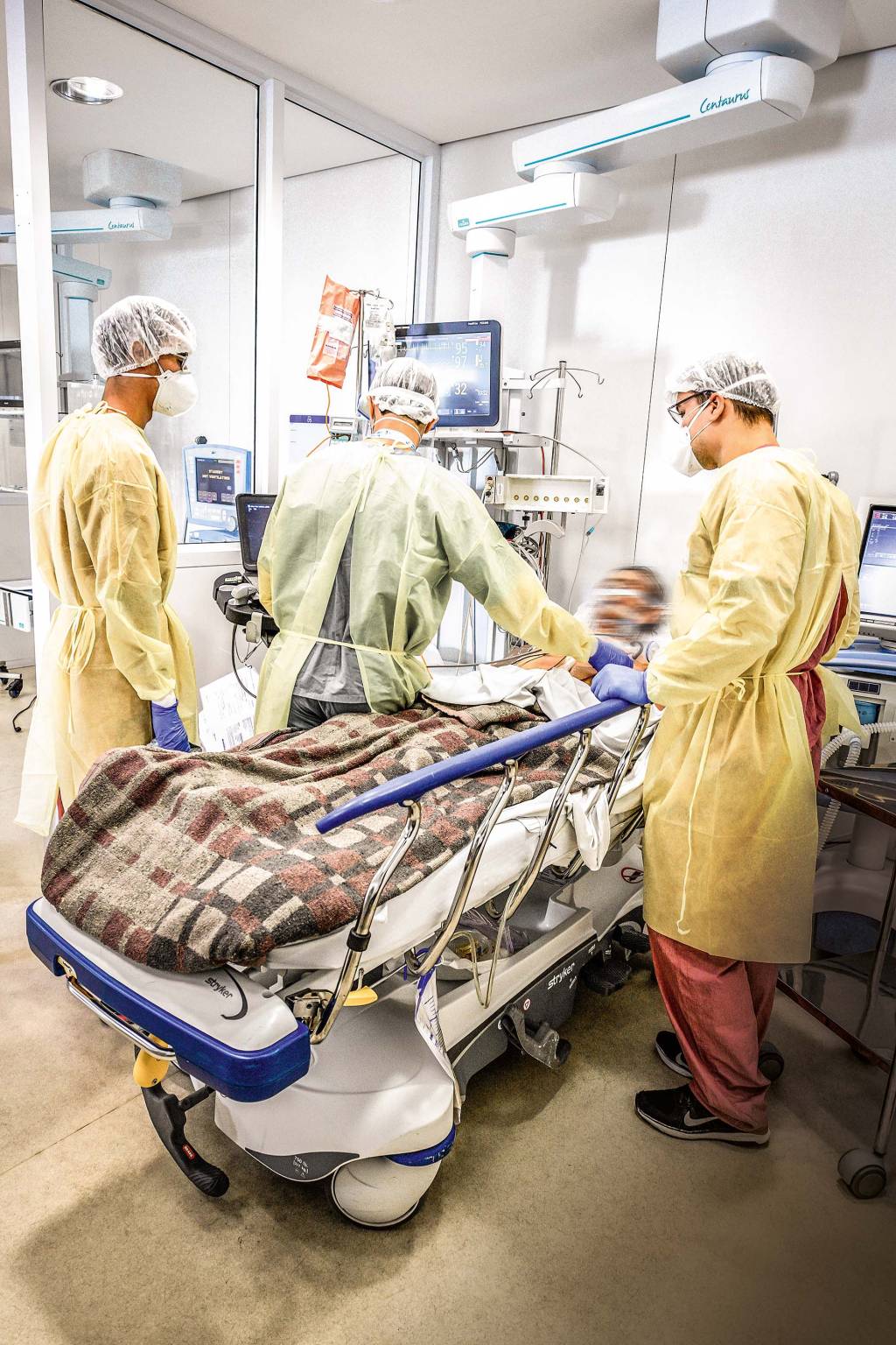 A imagem mostra um paciente deitado em uma cama de UTI cercado por três médicos que o observam. Todos estão com os rostos borrados.