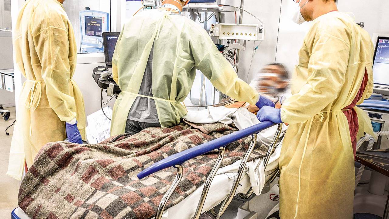 A imagem mostra um paciente deitado em uma cama de UTI cercado por três médicos que o observam. Todos estão com os rostos borrados.