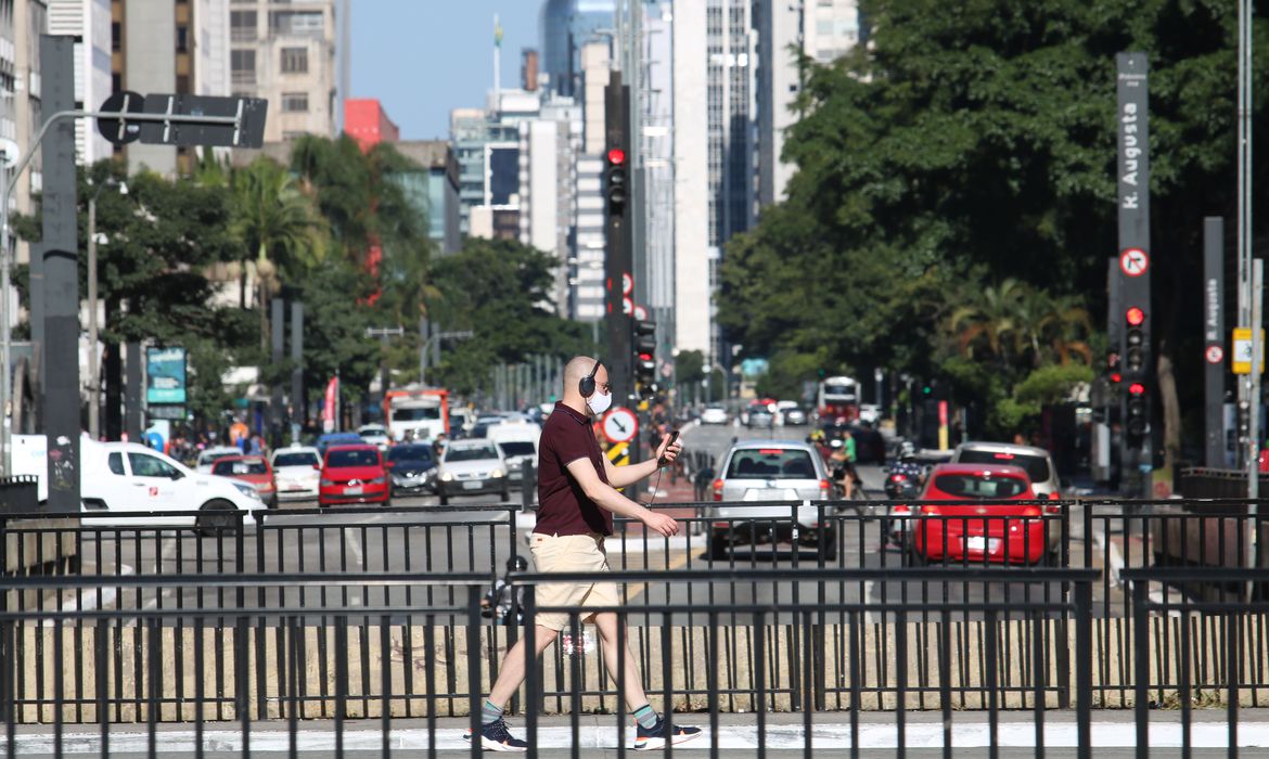 Imagem mostra pedestre caminhando de máscara e fones de ouvido na Avenida Paulista
