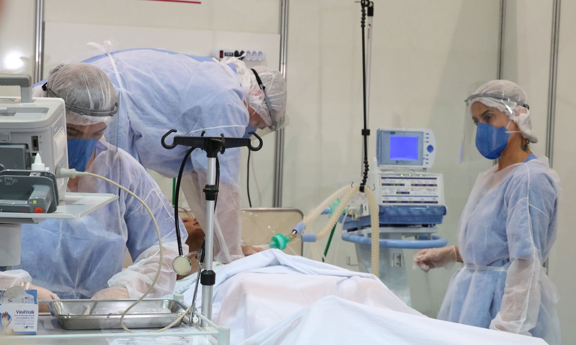 Imagem mostra ambiente hospitalar, com equipamentos, enfermeira e paciente deitado em cama