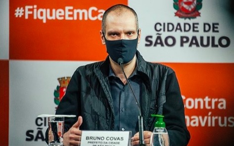 Bruno Covas aparece sentando em bancada durante coletiva de imprensa, usando máscara, com logotipo da prefeitura ao fundo