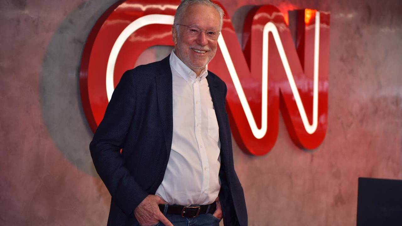 A imagem apresenta Garcia, em frente ao logo da CNN, com uma roupa social e sorrindo para a câmera